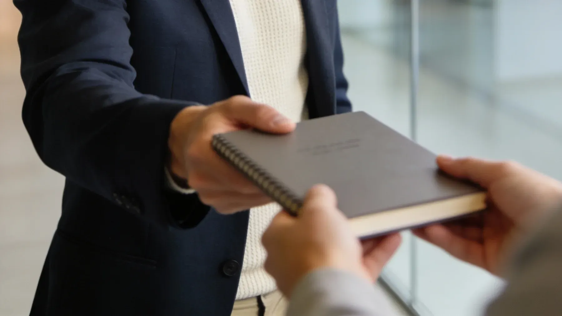 Two people exchange a notebook with both sets of hands still attached, turning a shared object into a decision about possession, memory, and obligation.