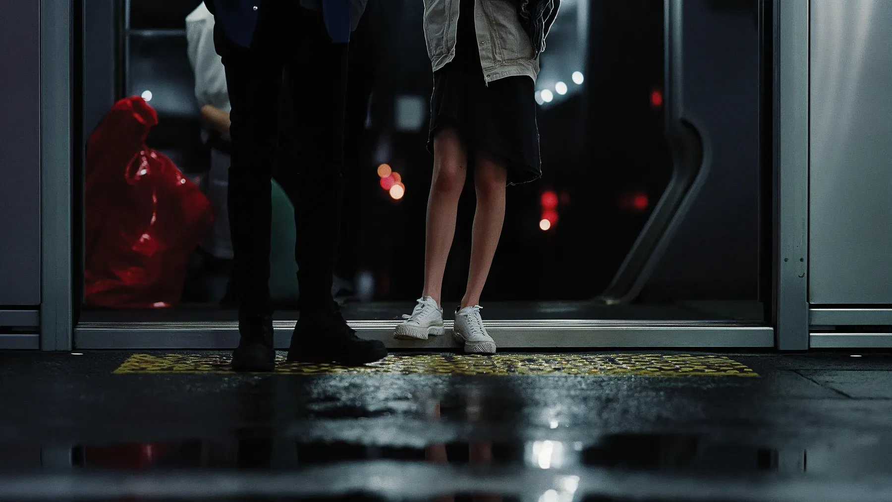 Two people pause at the threshold of a train on a wet night, where the opportunity for one more exchange is visibly running out.