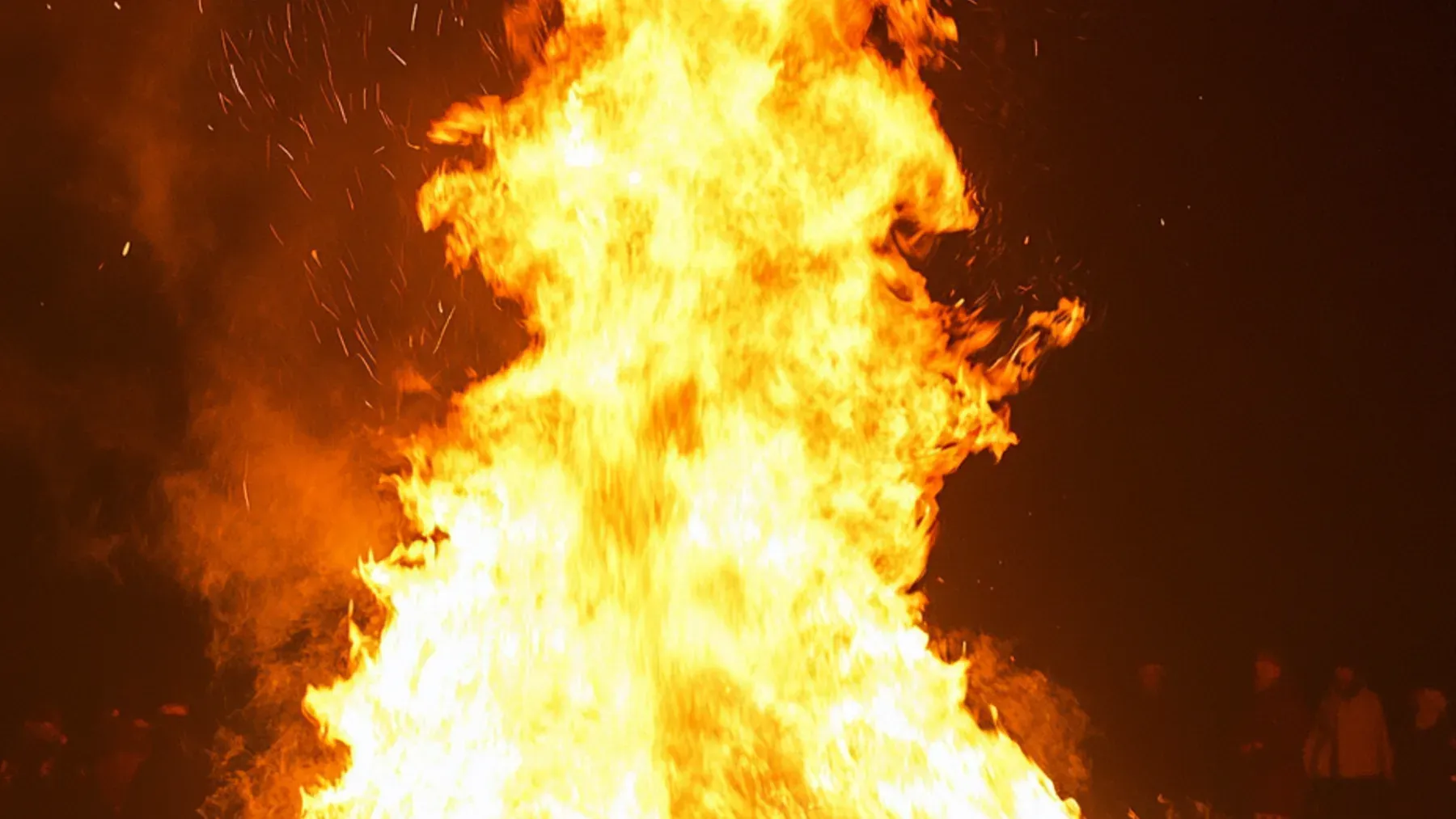 A bonfire rising above figures at night, representing ritual as a public sequence that distributes roles, attention, memory, and authority.