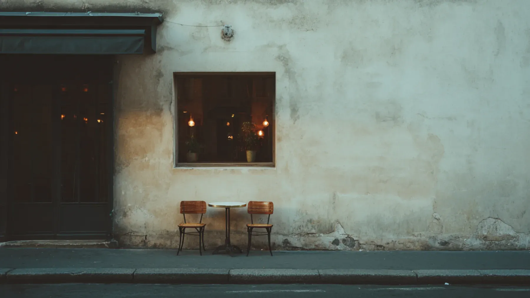 Two empty cafe chairs wait beside a small table, preserving the outline of a shared ritual after neither friend arrives to occupy it.