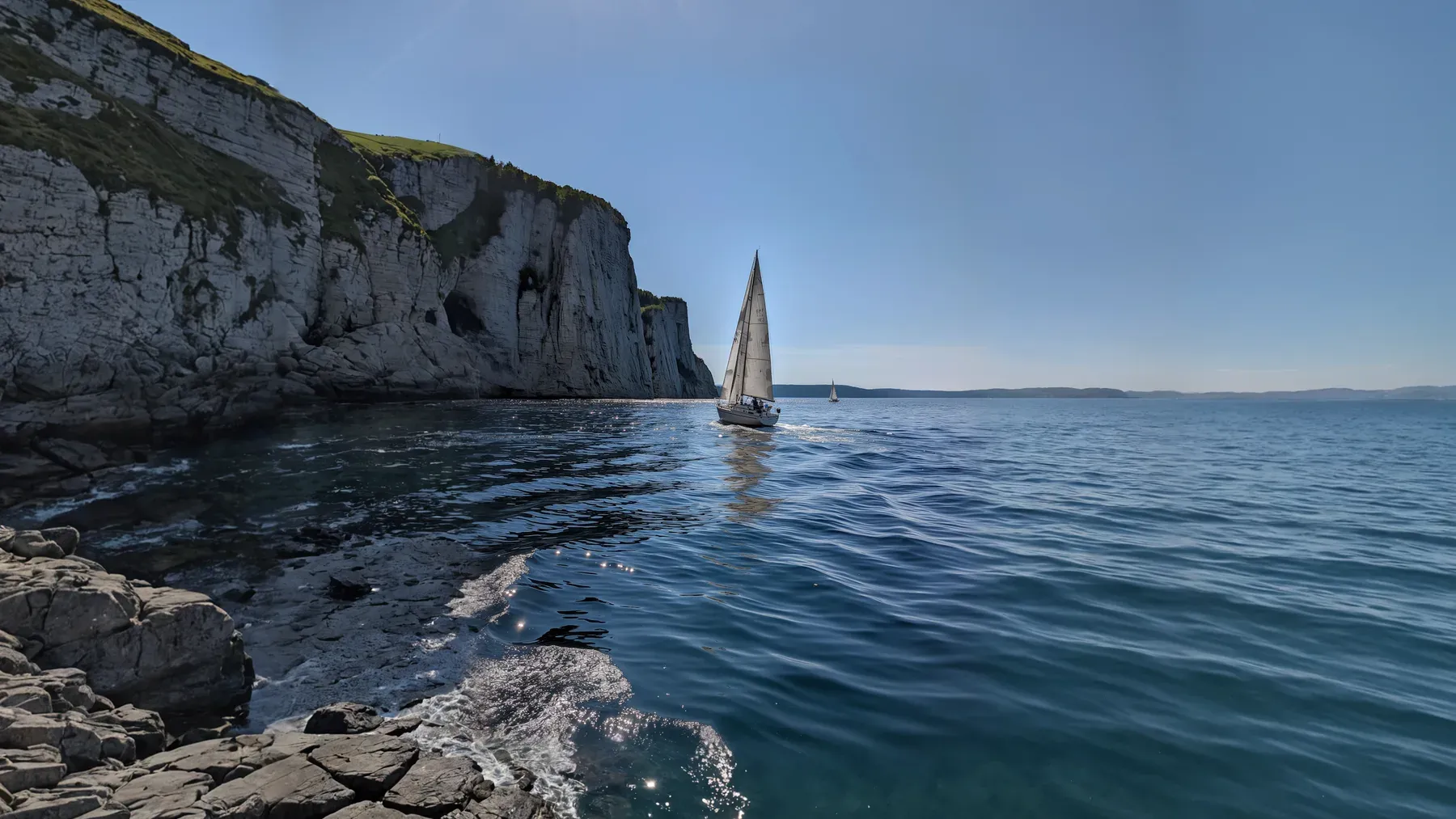 A lone sailboat passes a rocky coast, emphasizing the narrow relationship between route, geography, and a crew's shared decisions.