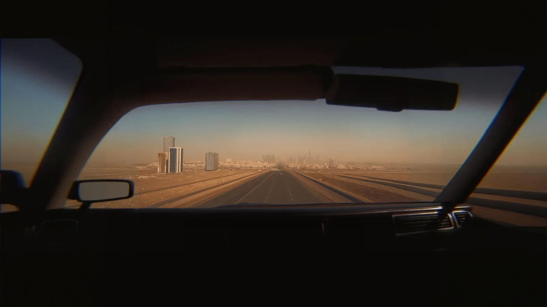 A highway crosses a dry landscape toward a distant city, showing a route that joins movement, destination, and the systems encountered between them.