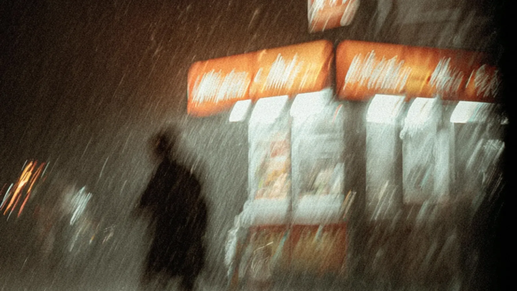 A figure crosses a rain-blurred city at night, showing the physical public world that bears the consequences of an invisible system decision.