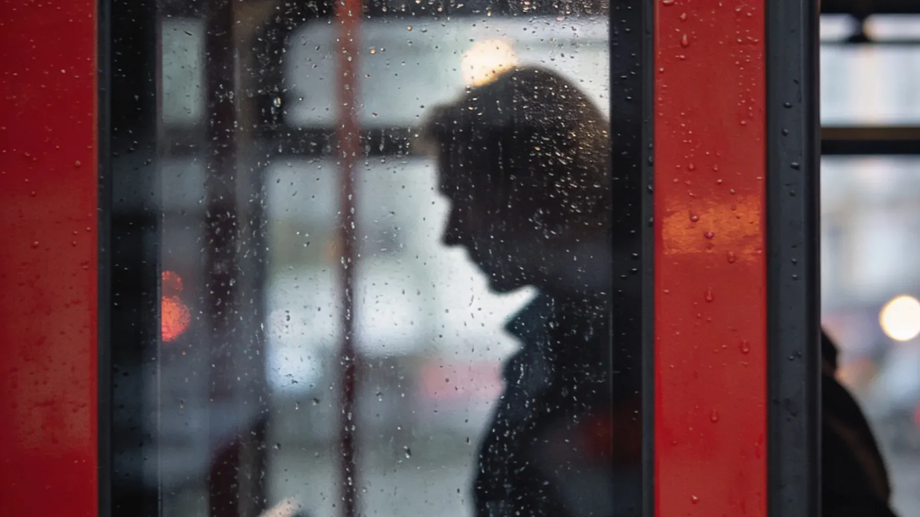 A commuter appears through a rain-covered transit window, where routine technology and supernatural evidence overlap.